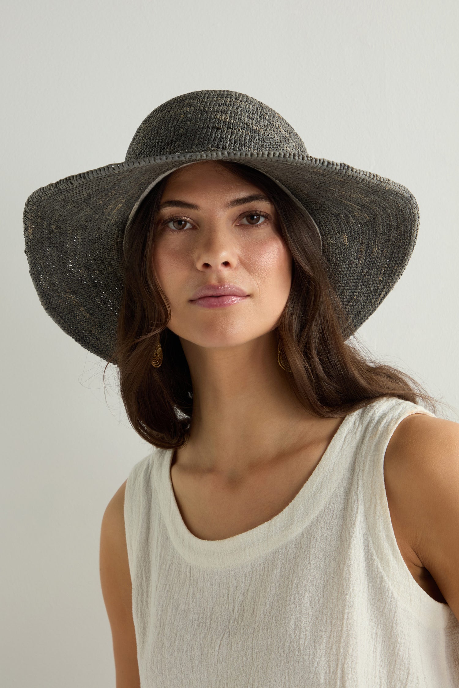 A person with long hair, wearing the Chapeau Westilux, a large raffia hat, and a sleeveless white top looks forward with a neutral expression.