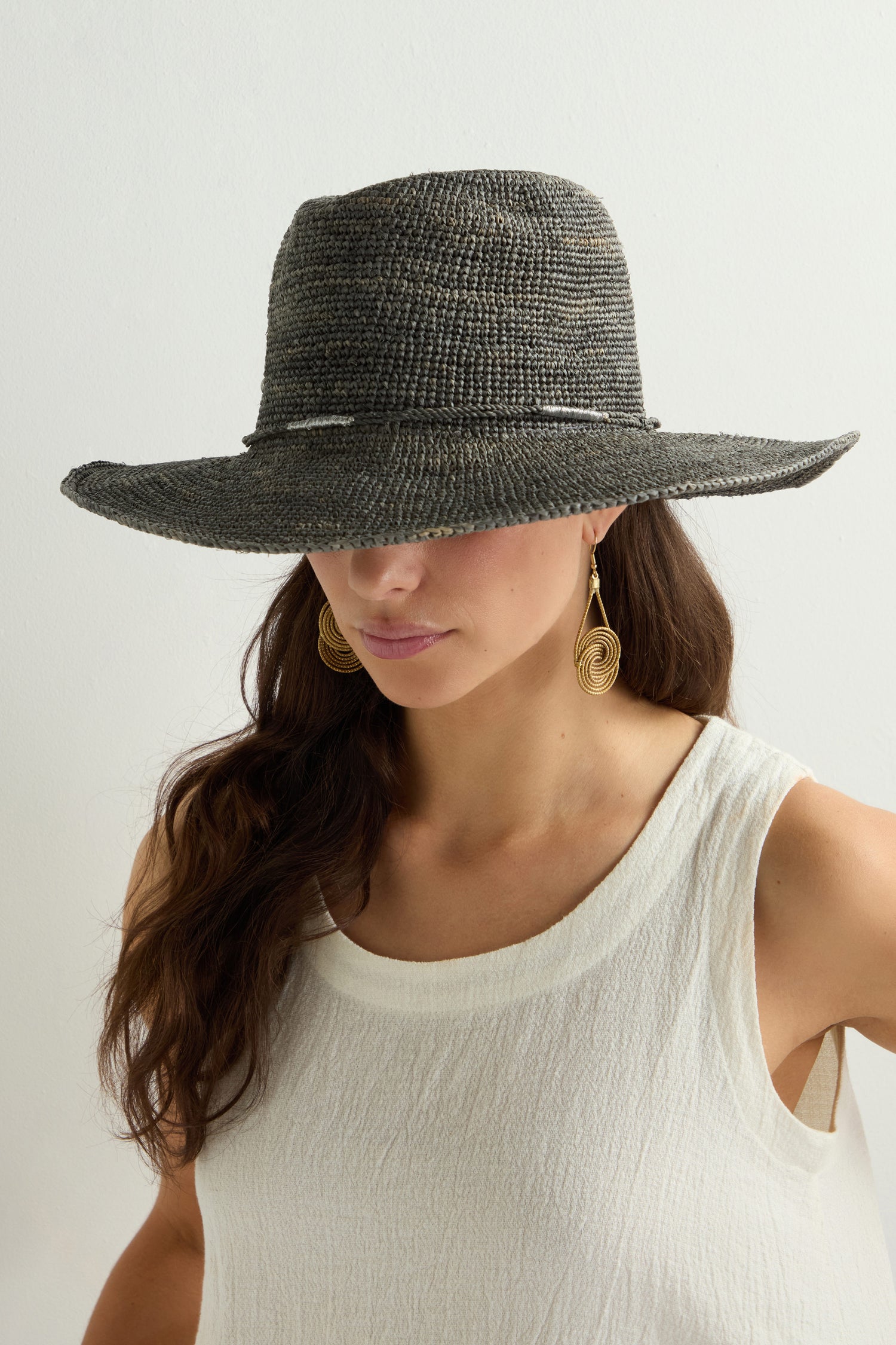 In a stylish Chapeau Westilux, its wide brim casting shade over her face, the woman wears gold hoop earrings and a white sleeveless top, embodying the essence of a breezy summer wardrobe.