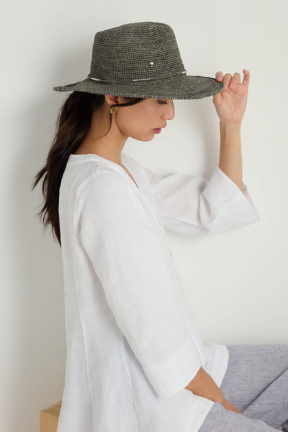 A woman in a white blouse and gray pants sits sideways, holding the brim of the Chapeau Westilux handwoven raffia summer hat, set against a plain light background.