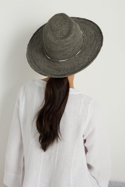 A person with long dark hair, wearing the Chapeau Westilux—a wide-brimmed handwoven raffia summer hat—and a white shirt, faces away from the camera against a plain background.