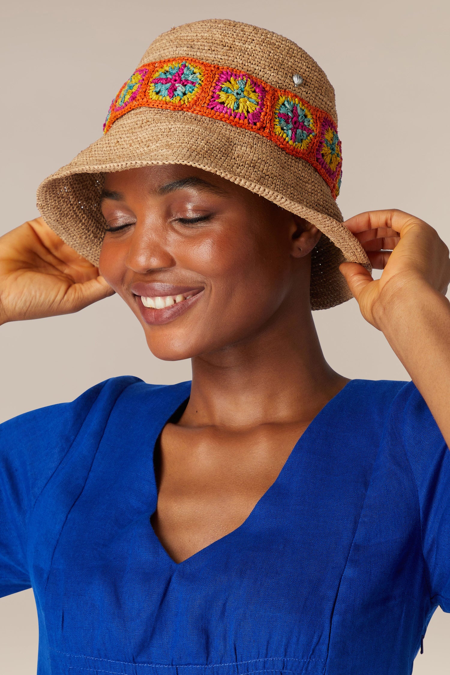 A person wearing a blue dress and a Chapeau Capizoa with colorful square patterns smiles with eyes closed, holding the hat with both hands.