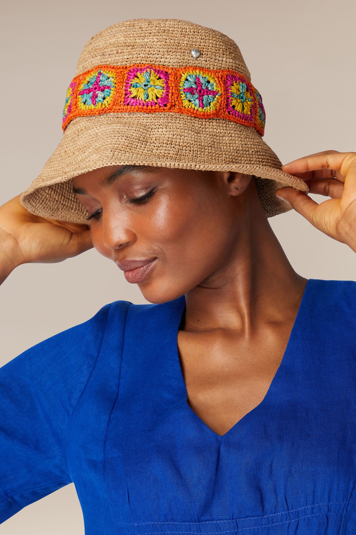 A person wearing a blue shirt and a Chapeau Capizoa with colorful crochet squares adjusts the hat with their hands.