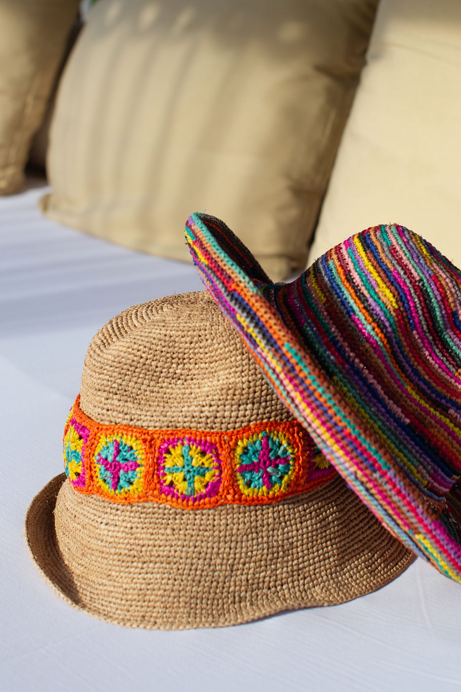 Two woven straw hats on a white surface; one, a Chapeau Capizoa, features a colorful band with geometric patterns, and the other has multicolored stripes throughout. Beige cushions are blurred in the background—perfect travel companions for sunny adventures.