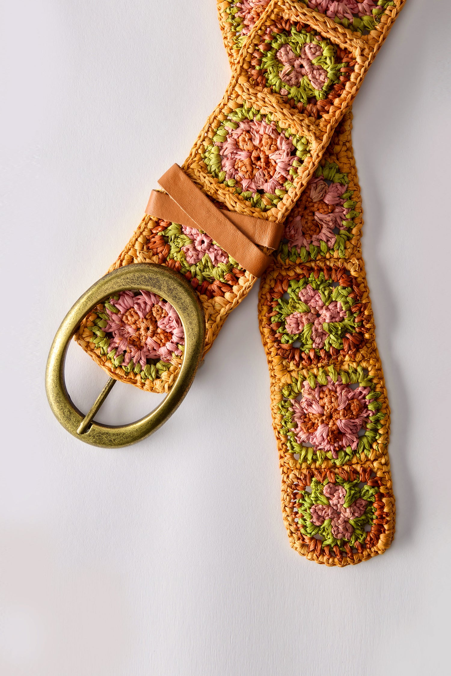 The Cinzoa Raffia Belt is a boho-chic accessory featuring a brass oval buckle and crafted in crochet-style raffia with a floral pattern in pink, green, and orange.