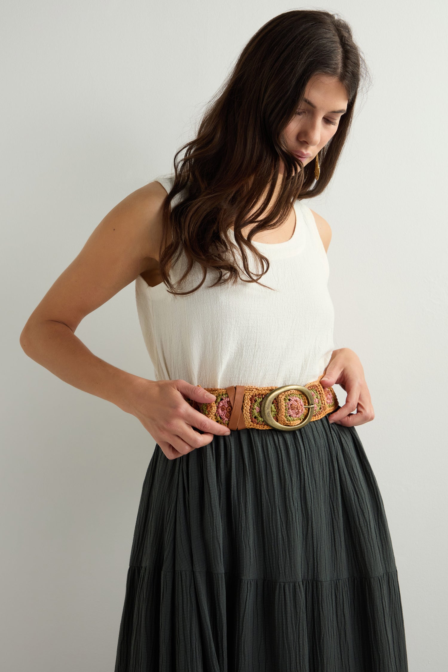 A woman, standing against a plain background, gazes downward while adjusting a vibrant Cinzoa Raffia Belt over her white sleeveless top and long dark skirt, adding flair to her look.