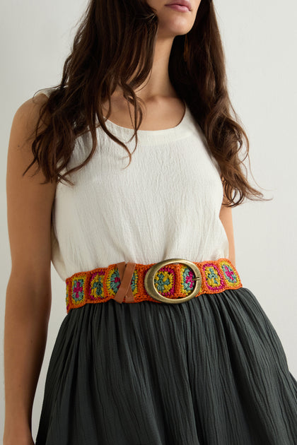 A woman in a sleeveless white top and dark skirt displays her boho-chic style with a colorful Rainbow Cinzoa Raffia Belt, featuring a large silver buckle, against a plain background.