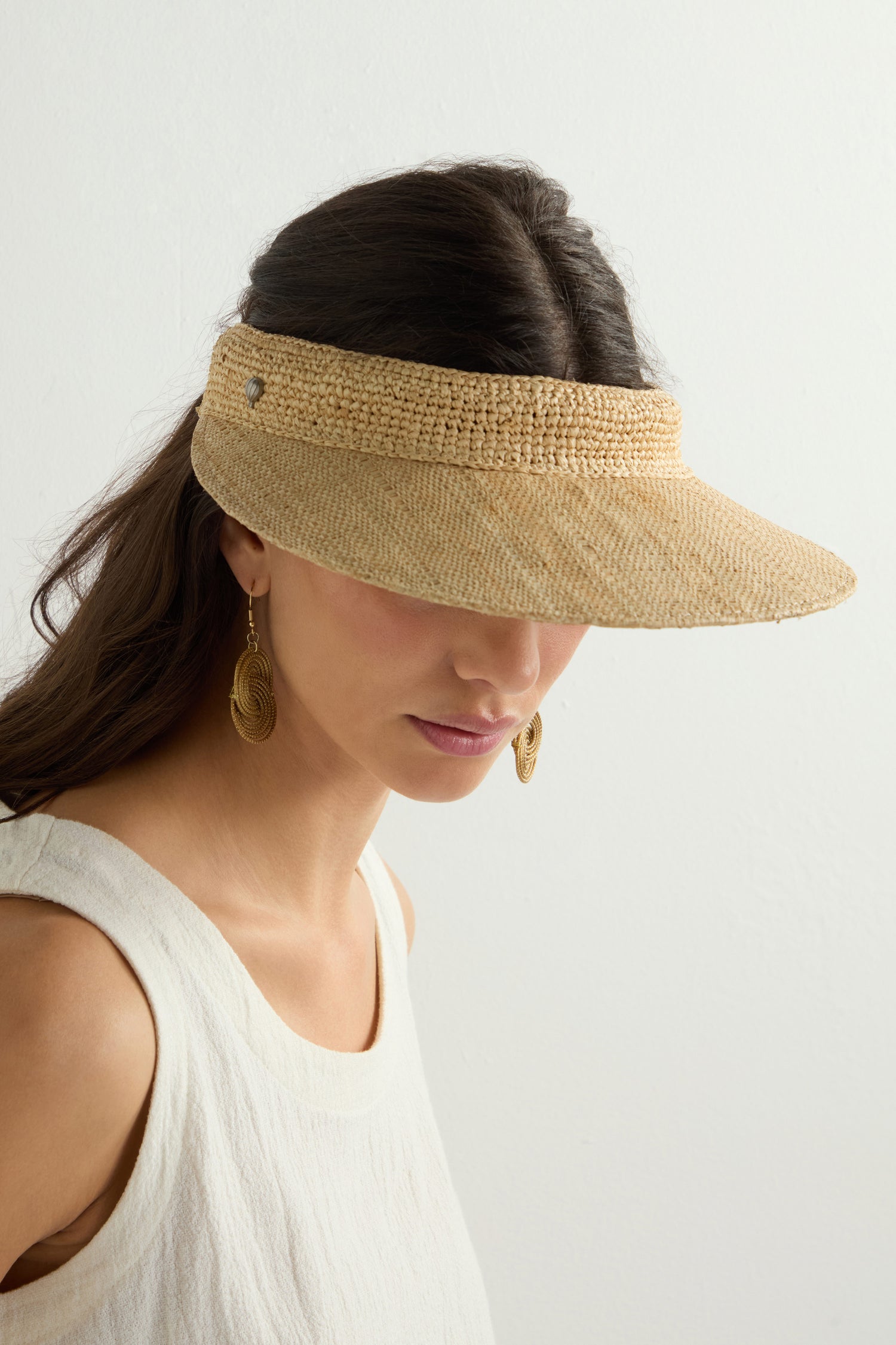 A woman with long dark hair wears a Garossoa Visor, highlighting elegant gold earrings as she faces slightly to the side against a plain backdrop, adding chic style and sun protection.