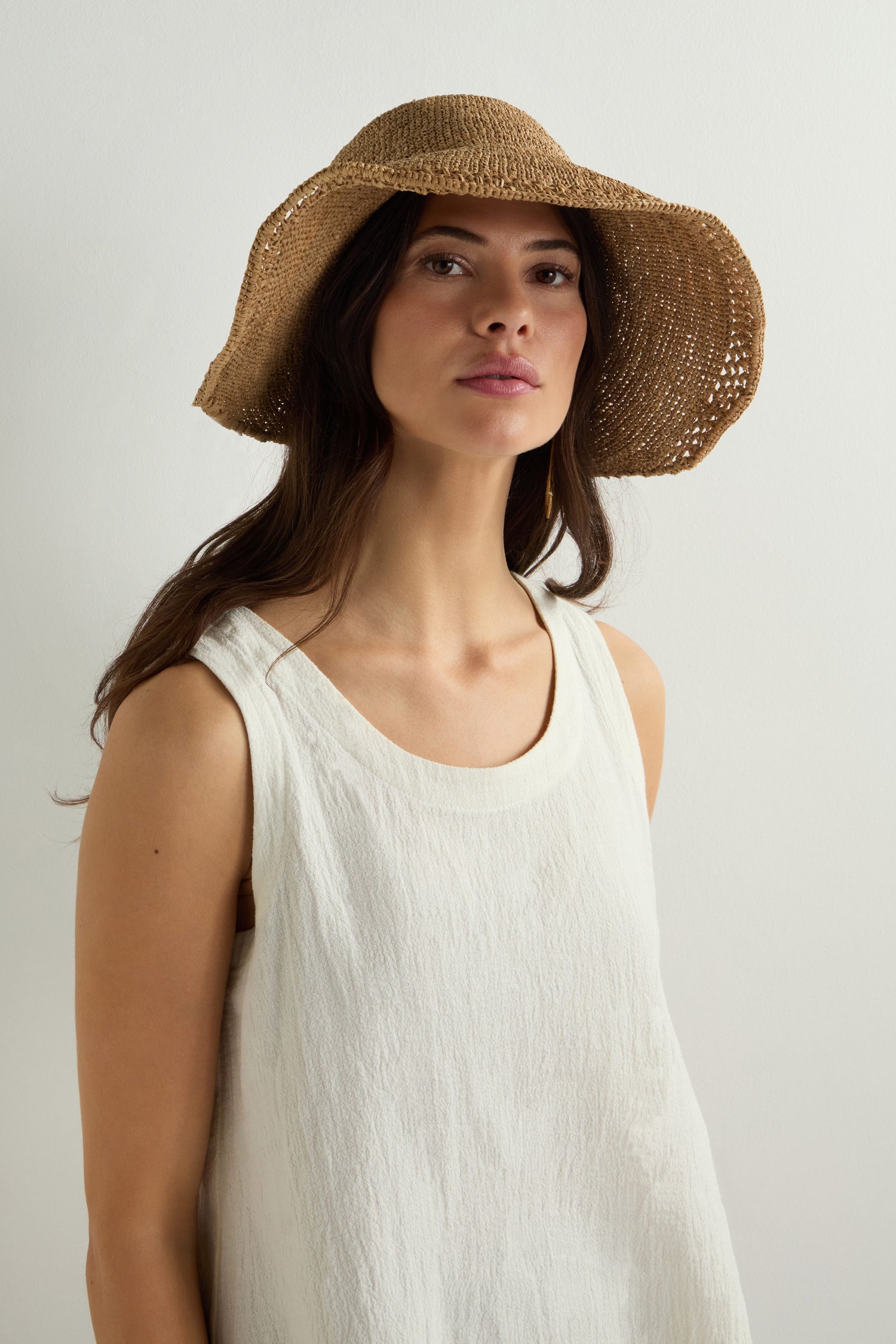 Wearing a white sleeveless top and the Chapeau Noloa, a natural woven straw sun hat, against a neutral background perfectly captures the essence of summer style.