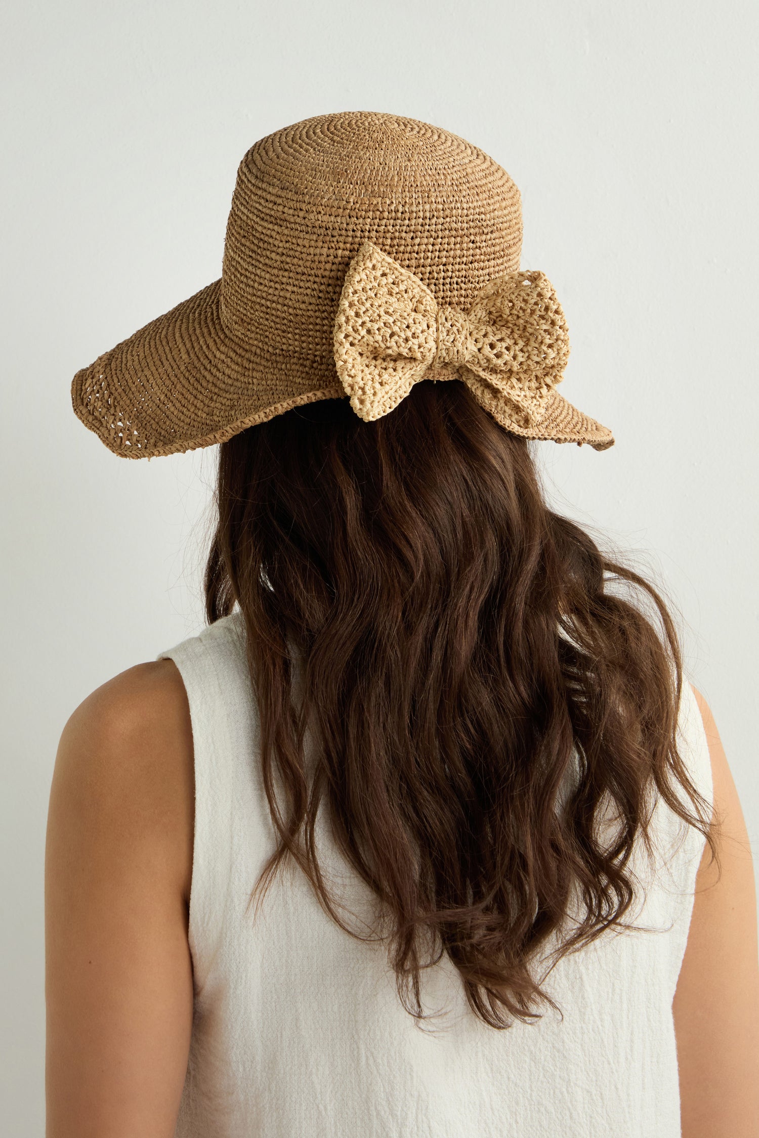 A person with long brown hair is seen from the back, wearing the Chapeau Noloa, a natural woven straw hat adorned with a large bow, epitomizing effortless summer style against a plain white background.