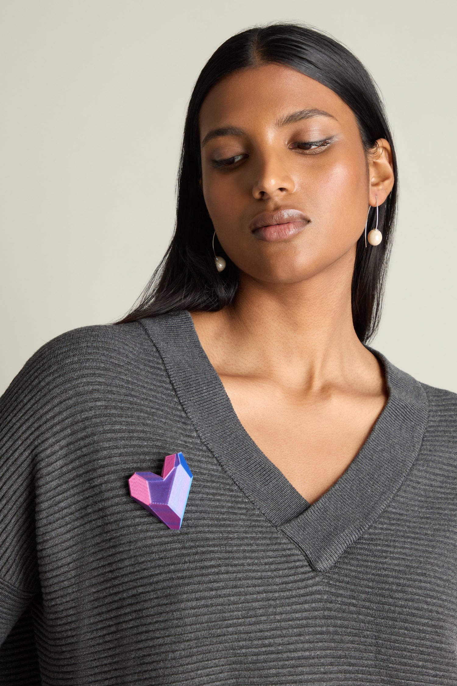 A person wearing a gray sweater and pearl earrings is looking down, showcasing modern accessory design with the vibrant, geometric 3D Printed Heart Brooch pinned to their sweater.