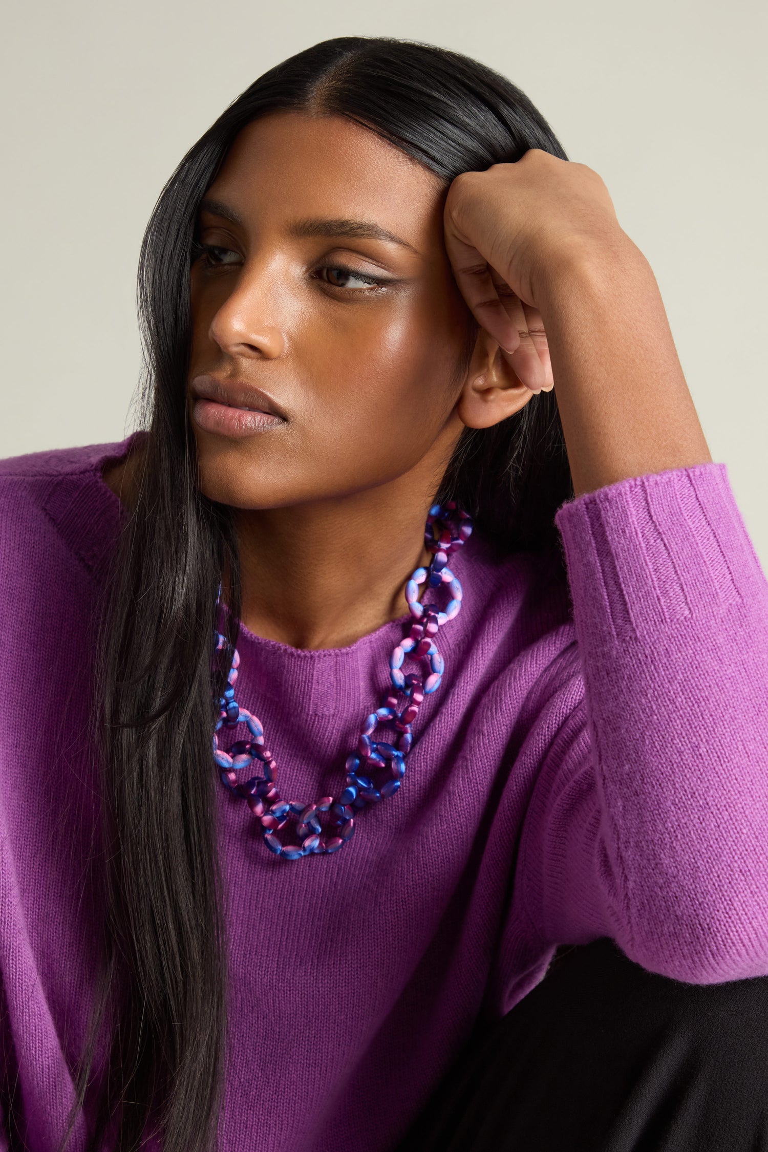 A woman with long black hair, dressed in a purple sweater and wearing a Maison 203 3D Printed Links Necklace, rests her head on her hand as she gazes thoughtfully to the side. The multicolored necklace is made with 3D printed links, offering an ultra-lightweight design that perfectly complements her pensive demeanor.