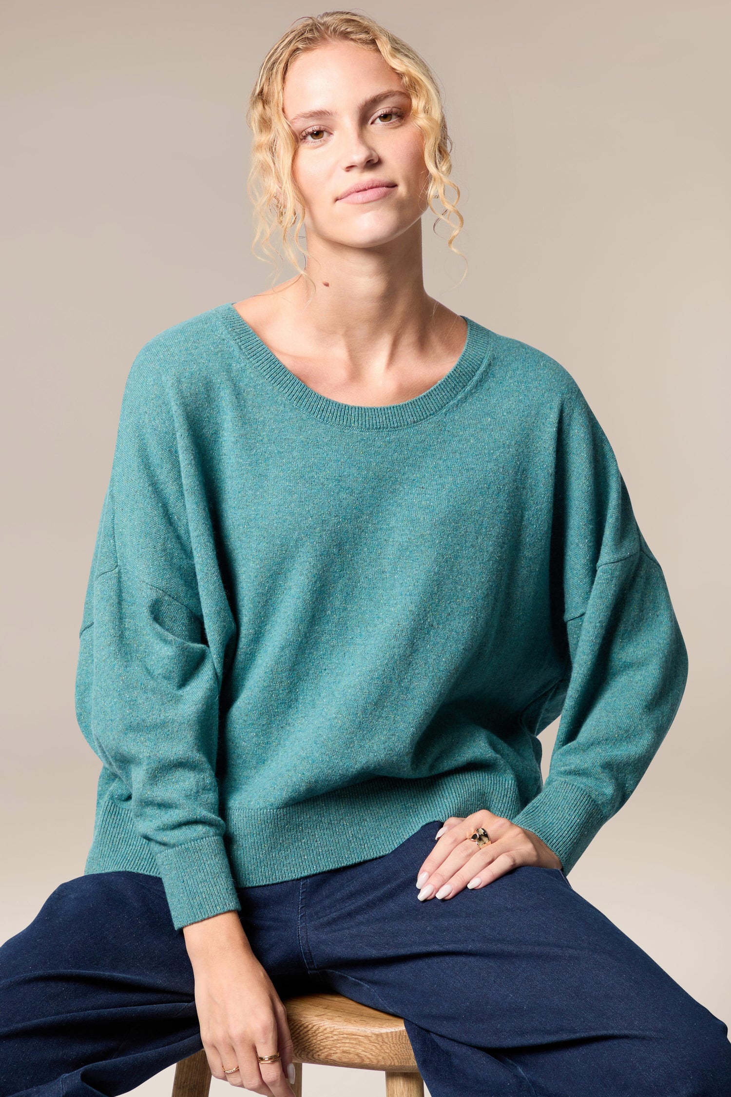 A woman is sitting on a stool wearing the Granit Scoop Knit teal sweater.