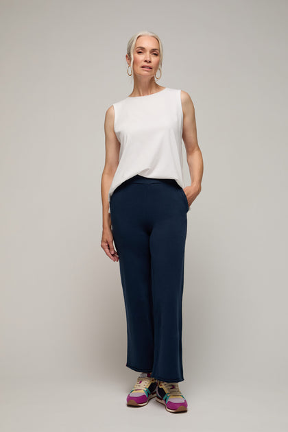 A woman with gray hair stands against a neutral background, wearing a white sleeveless top, Lana Pant in navy blue with an elasticated waist, and colorful sneakers—ideal for elevated lounging.
