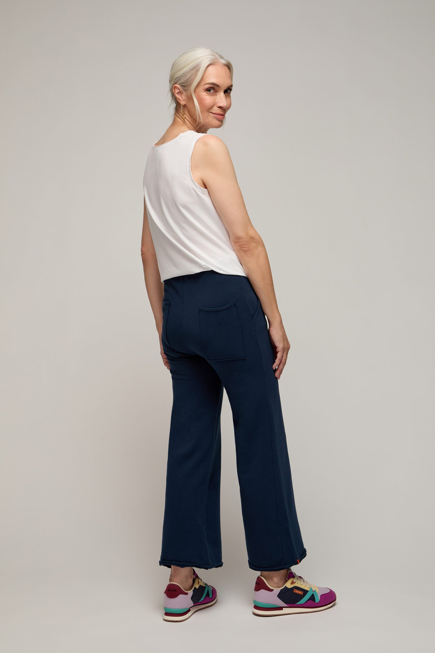 A woman with gray hair wears a white sleeveless top, the navy blue Lana Pant with an elastic waistband, and colorful sneakers as she stands looking over her shoulder—an effortlessly chic look for elevated lounging.