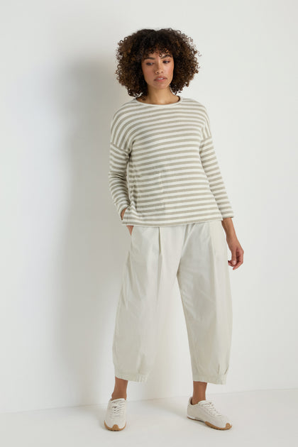 A person stands against a plain white wall, wearing a beige and white striped long-sleeve top, Pleated Poplin Trousers with a tapered leg, and white sneakers.