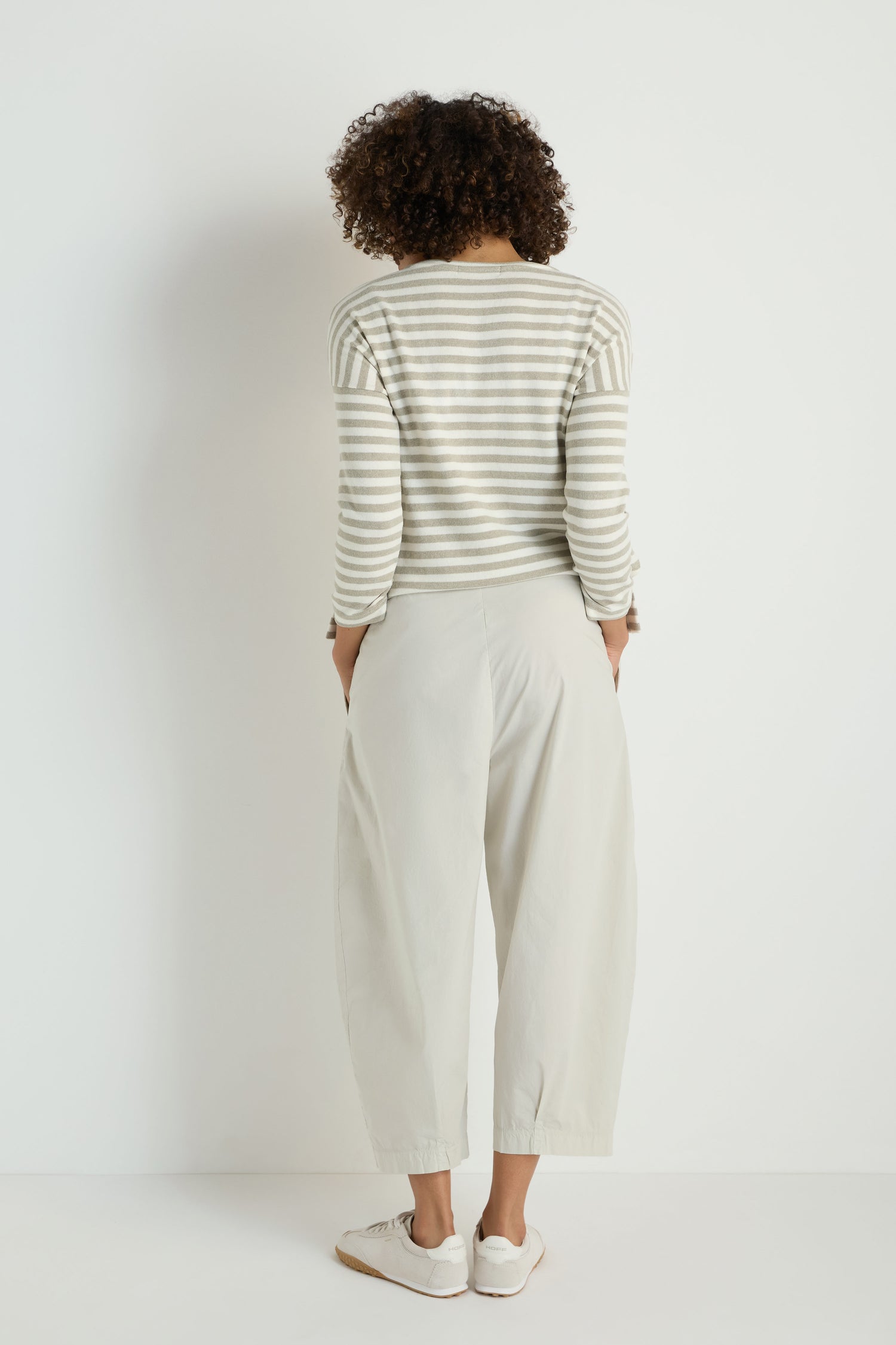 A person stands with their back to the camera, wearing a gray and white striped long-sleeve shirt, Pleated Poplin Trousers, and white sneakers against a plain white background.