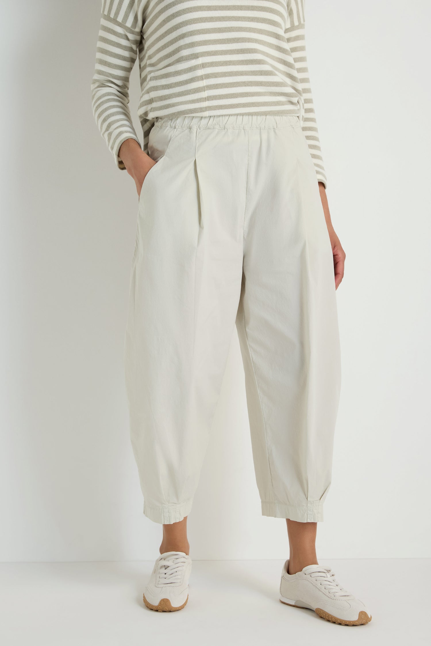 A person stands against a plain background wearing a beige and white striped long-sleeve shirt, Pleated Poplin Trousers with a tapered leg in a cotton blend poplin, and white sneakers.