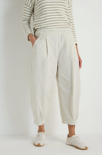 A person stands against a plain background wearing a beige and white striped long-sleeve shirt, Pleated Poplin Trousers with a tapered leg in a cotton blend poplin, and white sneakers.