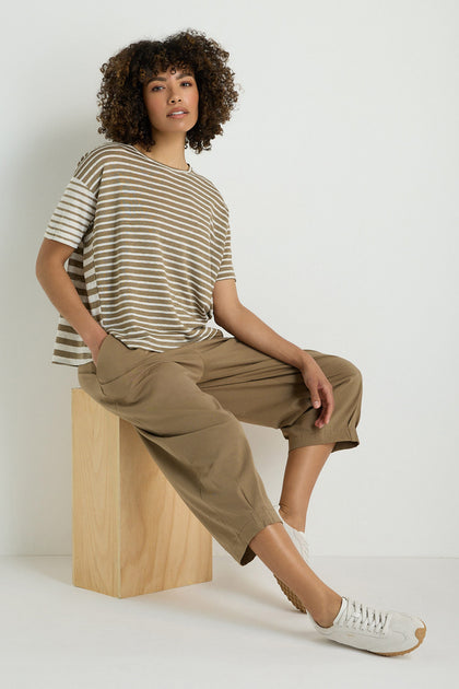 A person wearing a striped short-sleeve shirt, Pleated Jersey Trousers with an elasticated waist, and light sneakers sits on a wooden block against a plain white background.