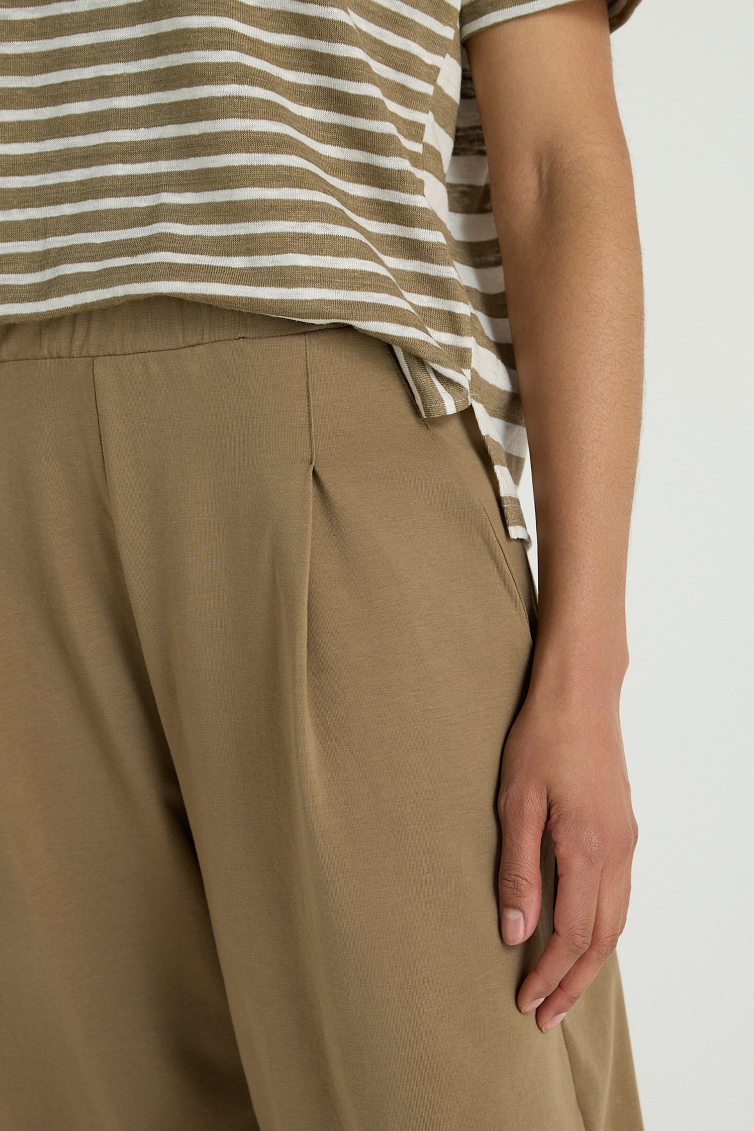 A person wears a beige and white striped shirt paired with Pleated Jersey Trousers featuring an elasticated waist, their right hand resting by their side.