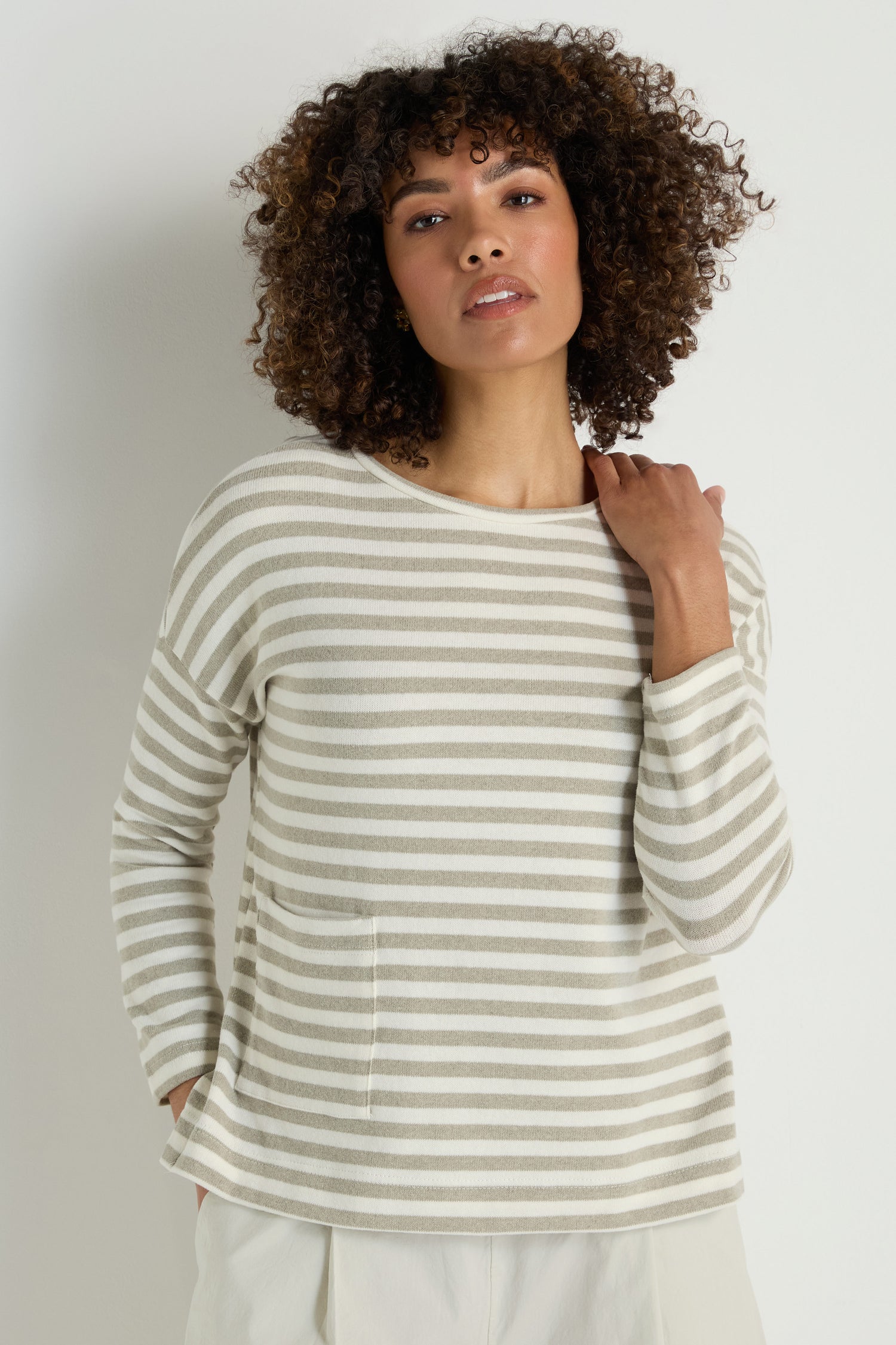 A woman with curly hair wears the Stripe Pocket Fleece Top in a relaxed beige and white stripe design with light pants, standing against a plain light background.