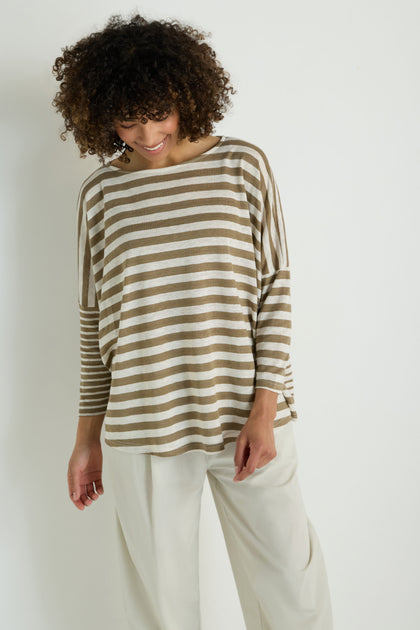 A woman stands against a plain background, looking down and smiling while wearing the Oversize Stripe Tunic in beige and white, paired with off-white pants.