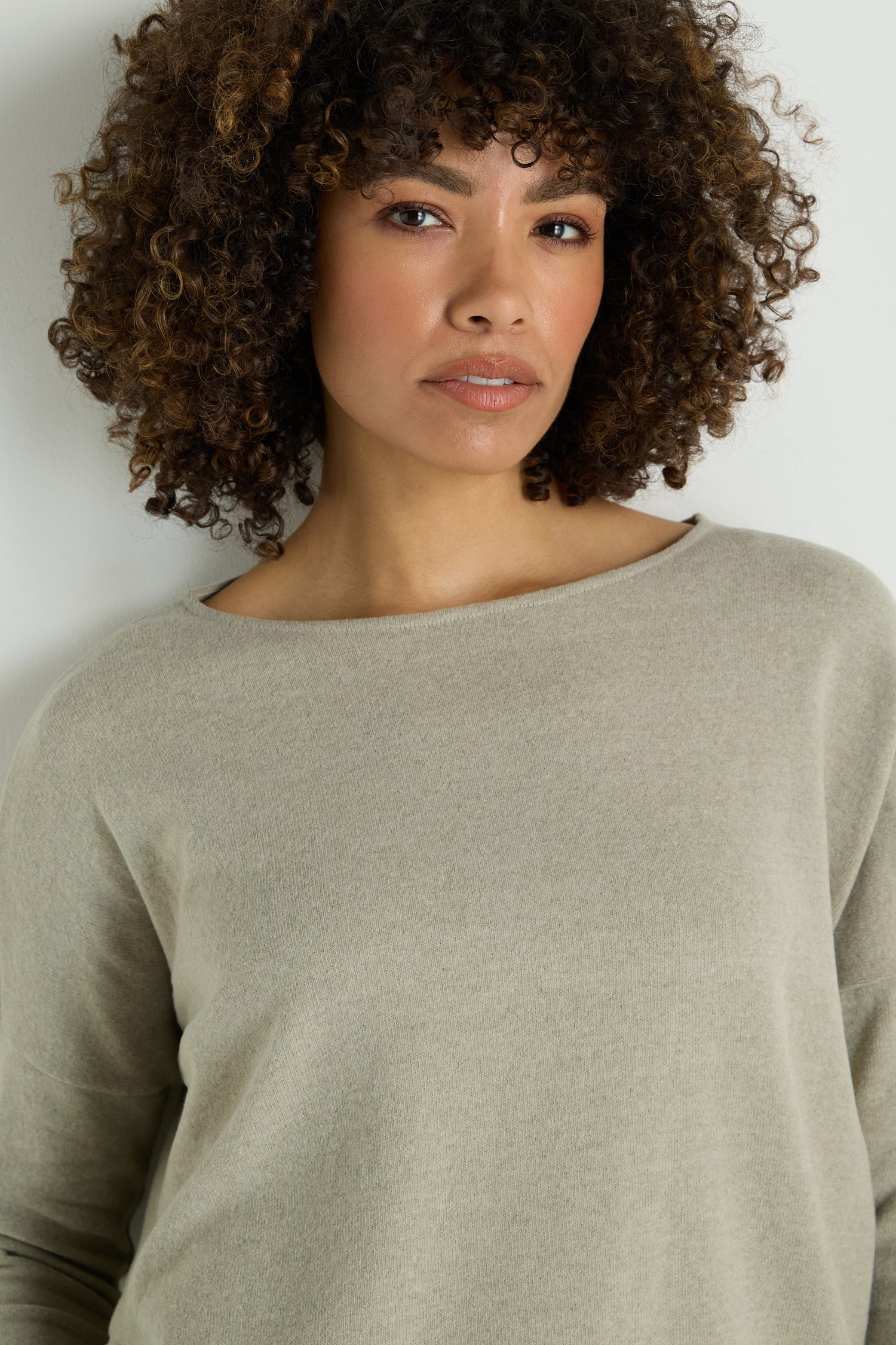 A woman with curly hair wears the Flared Fleece Top, a light beige cotton-blend long-sleeve, standing against a plain white background and looking at the camera with a neutral expression.
