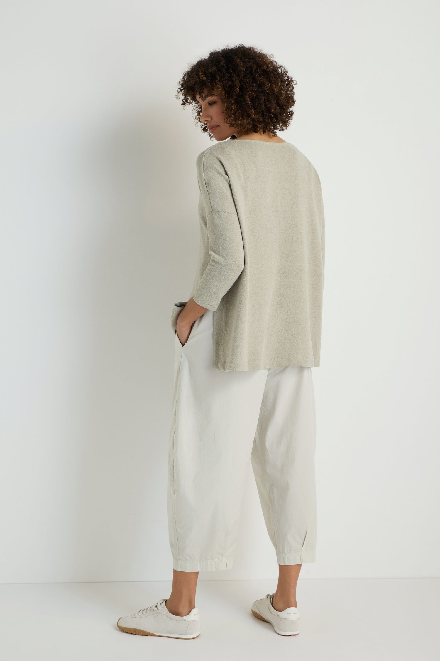 A person stands facing away, wearing a loose light gray Flared Fleece Top, wide-leg white pants, and white sneakers against a plain white background.