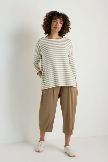A woman stands against a white wall wearing the Stripe Flared Fleece Top, styled with loose tan pants and white sneakers.