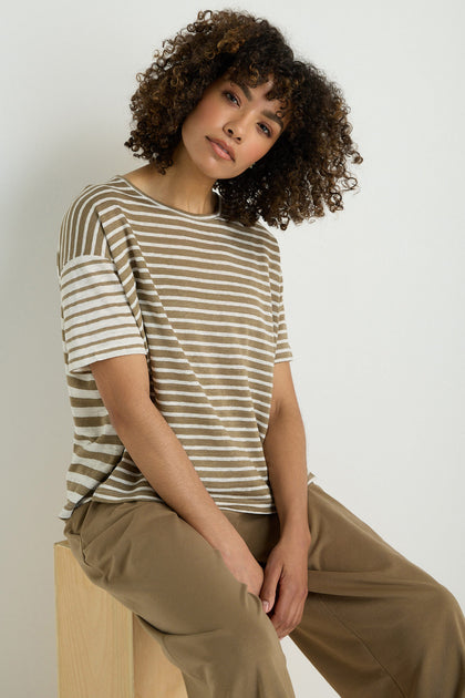 A woman with curly hair sits on a wooden block, wearing the Mixed Stripe T-Shirt—a beige and white linen-viscose blend—paired with tan pants, set against a plain light background.