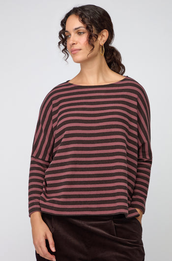 A woman stands slightly sideways, wearing a loose Martis Scoop Neck Wide Stripe Top with wide-leg dark trousers and black boots, set against a plain light background.