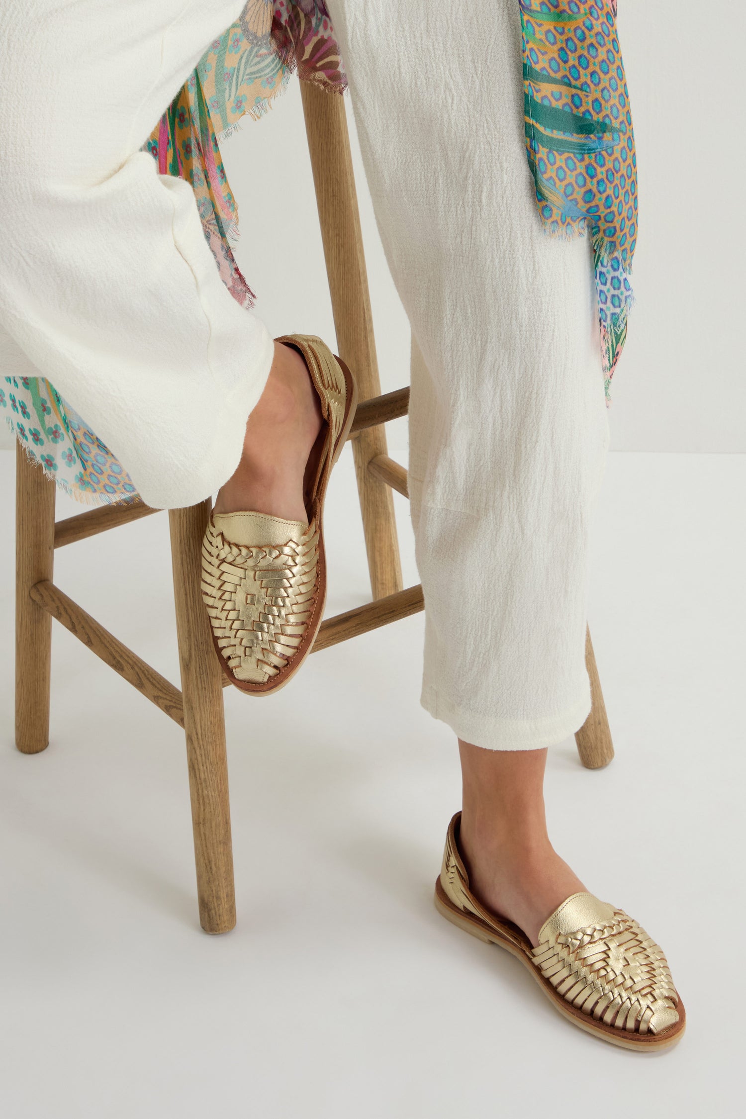 Sitting on a wooden stool, a person wearing white pants and gold slip-ons relishes the breathable comfort of their attire. The backdrop is vivid and patterned, echoing the vibrant designs associated with handcrafted Mexican goods, much like the Alegre Leather Huarache Sandals.