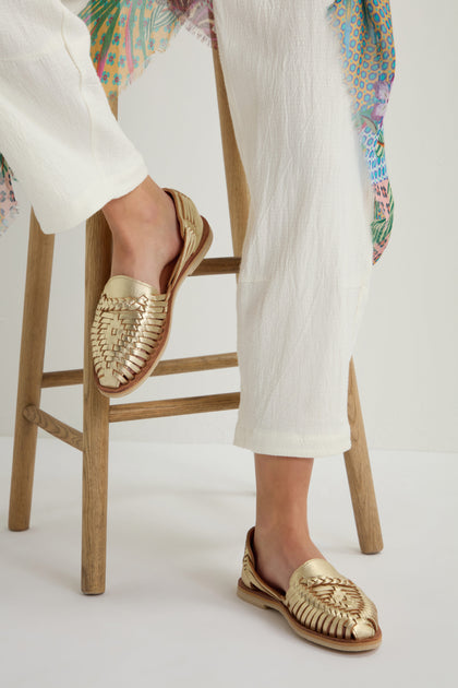 Seated on a wooden stool, a person wearing white pants and the handcrafted Alegre Leather Huarache Sandals from Mexico exudes breathable comfort.