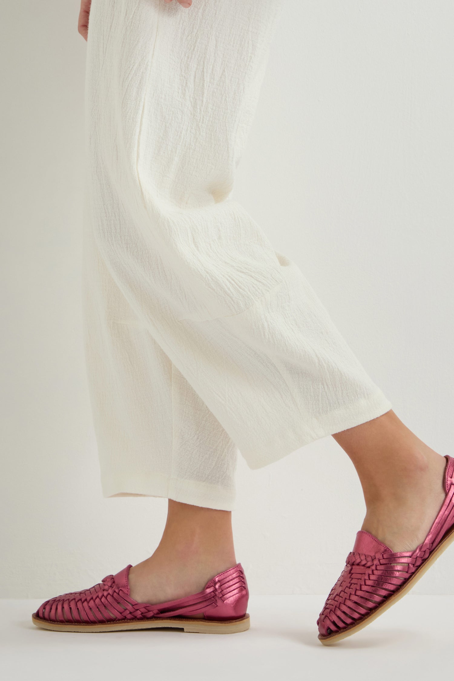 A person in cream pants and metallic pink shoes, reflecting the handcrafted style of Alegre Leather Huarache Sandals, stands against a plain background.