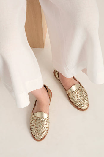 A person wearing white pants and Gold Alegre Leather Huarache Sandals, handcrafted in Mexico by Mapache Shoes, sits with their feet resting on a light-colored surface.