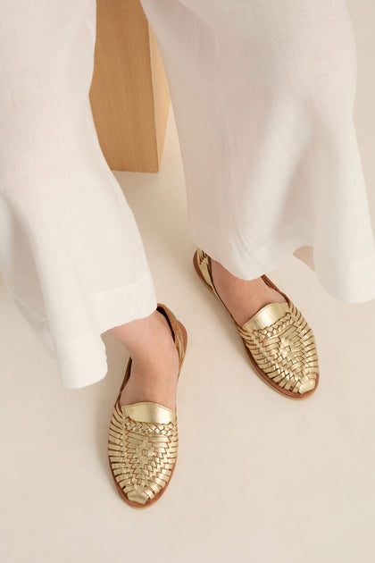 A person wearing white pants and Gold Alegre Leather Huarache Sandals, handcrafted in Mexico by Mapache Shoes, sits with their feet resting on a light-colored surface.