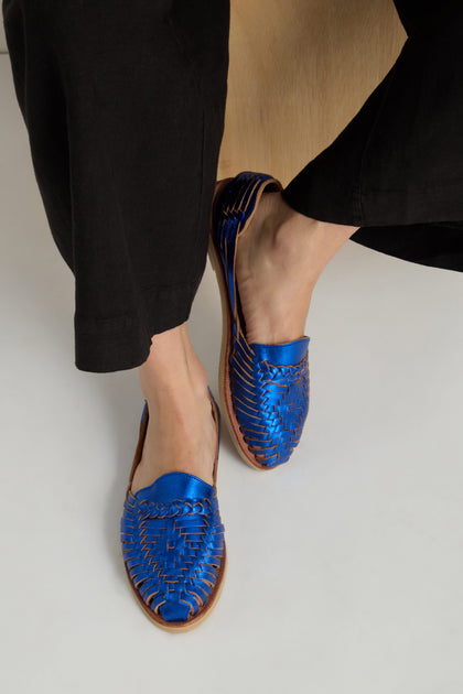 Wearing black pants and Metallic Blue Alegre Leather Huarache Sandals, handcrafted in Mexico with brown soles, standing on a light surface.