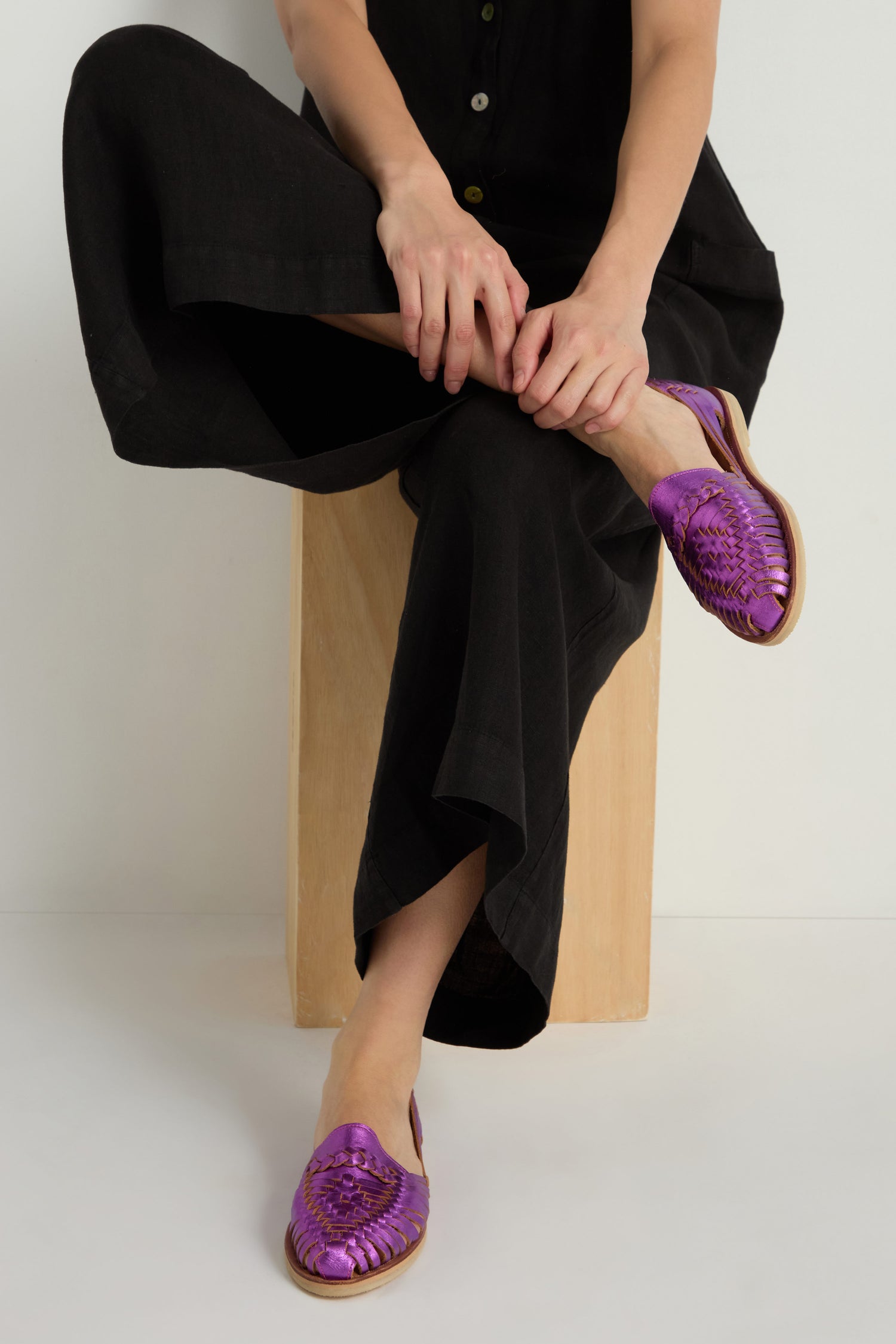 A person in black clothing sits cross-legged on a wooden block, wearing Metallic Purple Alegre Leather Huarache Sandals handcrafted in Mexico.
