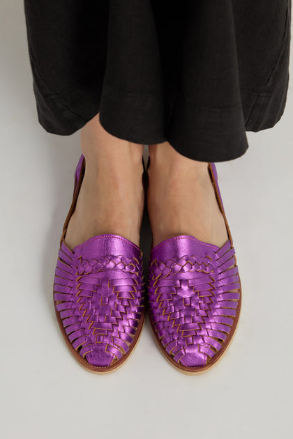 A person stands on a light surface wearing black pants and Metallic Purple Alegre Leather Huarache Sandals, handcrafted in Mexico.