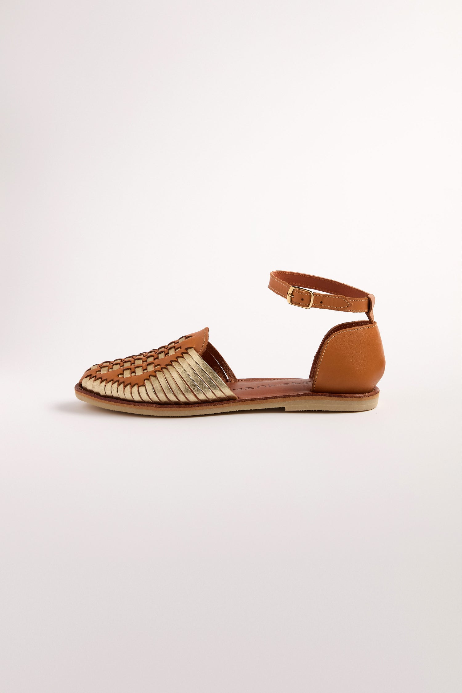 The Camila Two Tone Leather Sandals highlight their woven huarache design with a tan leather ankle strap and a brown heel. These handcrafted leather sandals have a flat sole, presented on a plain white background for an elegant look.