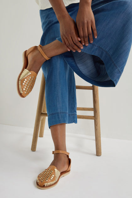 A person sits on a wooden stool wearing blue jeans and brown Camila Two Tone Leather Sandals with woven straps.