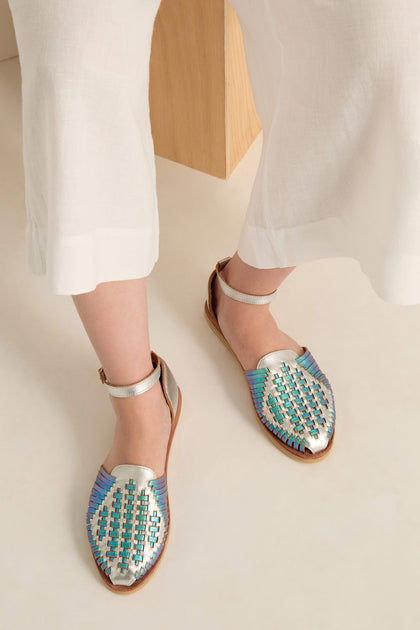 A person in white wide-leg pants wears Camila Mermaid Woven Sandals—iridescent with ankle straps—while seated on a light surface. This chic look features these online exclusive sandals.