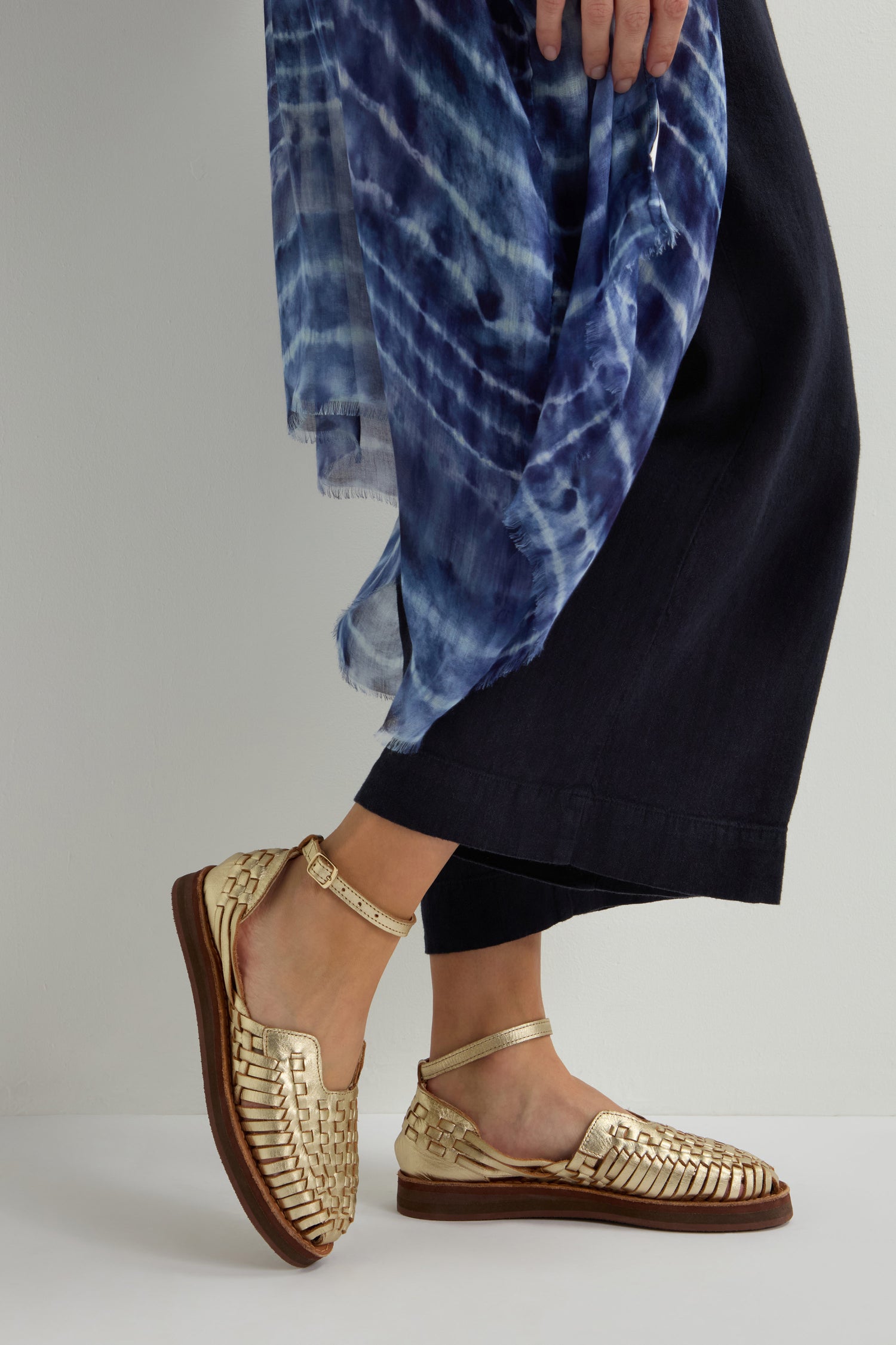 A person in Carmen Platform Woven Leather Sandals and black pants, holding a blue tie-dye scarf draped over their shoulder.