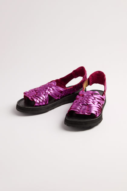 Bright pink Frida Woven Leather Sandals feature black soles, open toes, and ankle straps with a subtle metallic magenta finish. Handcrafted in Mexico, they are displayed on a white surface.