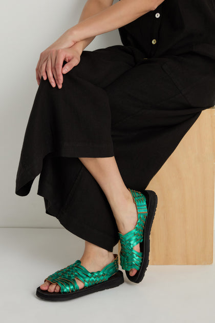 Seated on a wooden block with legs crossed and hands on knee, the person wears a black outfit and Frida Metallic Green Leather Sandals, handcrafted in Mexico.