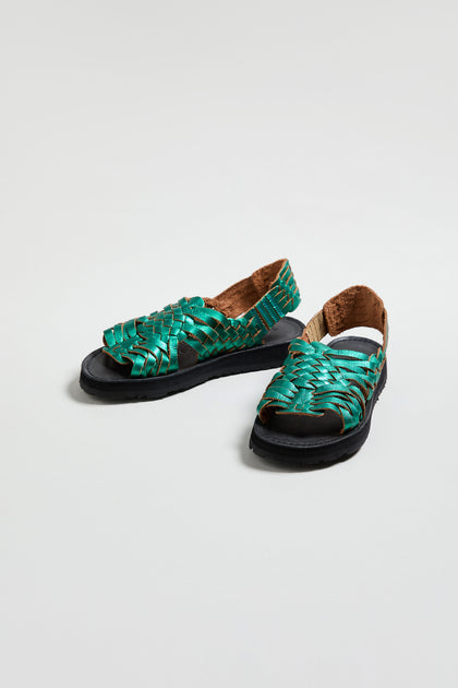 Frida Metallic Green Leather Sandals, handcrafted in Mexico, feature green woven leather straps, black soles, and a tan interior lining, displayed on a plain white background.
