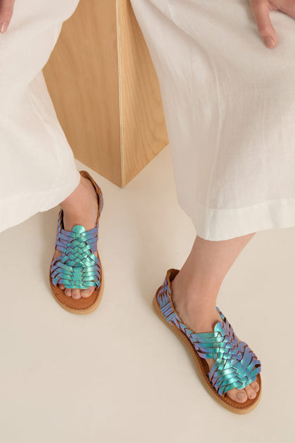 A person in white pants and Frida Mermaid Leather Sandals, handcrafted in Mexico from iridescent woven leather, sits on a wooden block with their feet resting on a light-colored floor.
