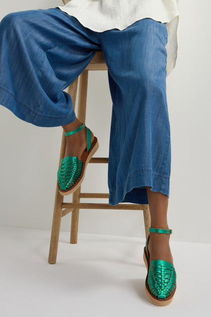 A person is seated on a wooden stool, wearing blue wide-legged pants and green Loma Platform Braided Leather Sandals with ankle straps.