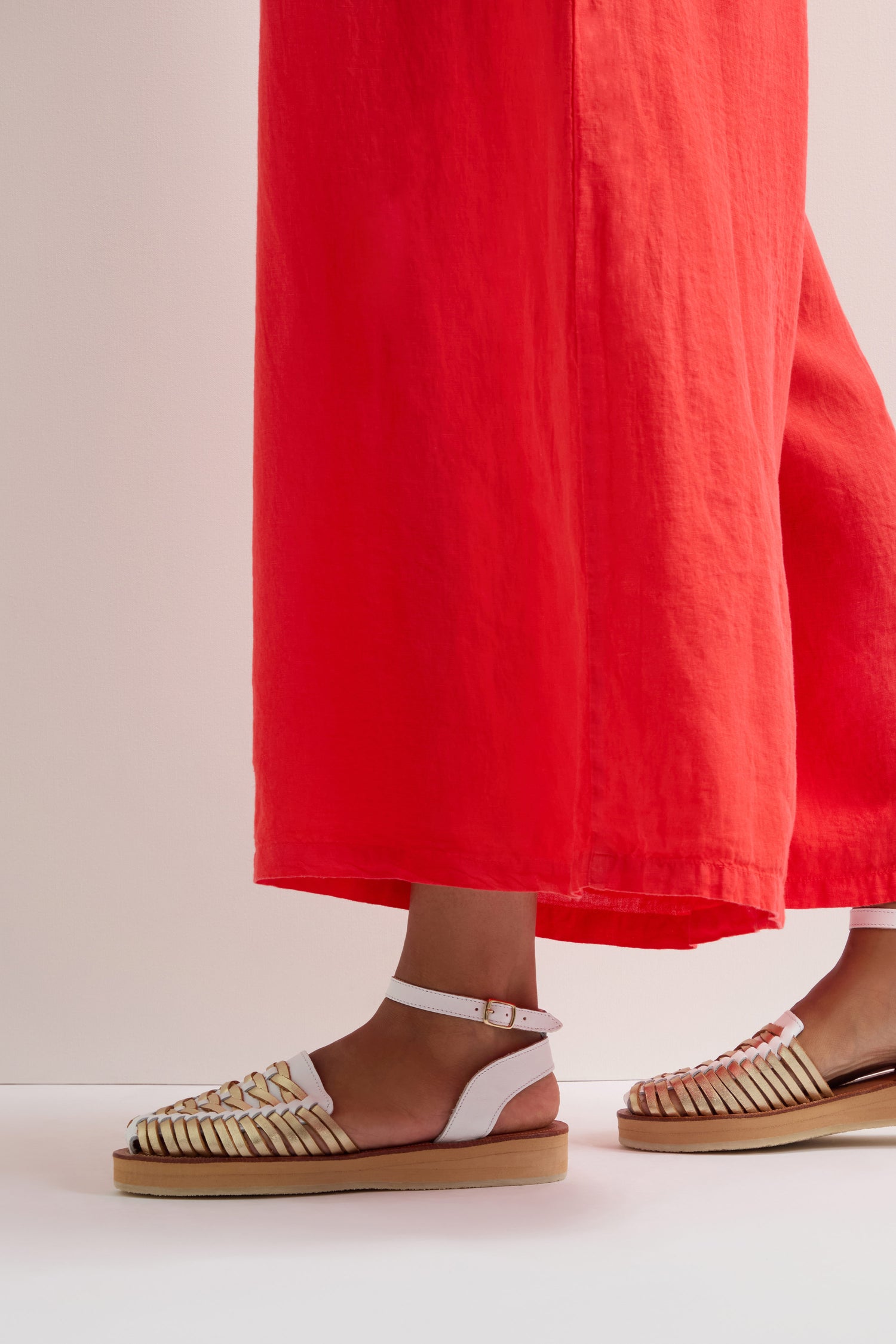 A person elegantly stands on a light-colored floor, wearing a long red skirt and Loma Platform Braided Leather Sandals. The white strappy design with a brown sole adds chic flair to the outfit.