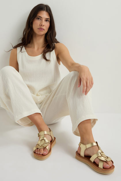 A woman in a white outfit sits against a wall, wearing elegant Tulum Leather Buckle Sandals with gold accents.